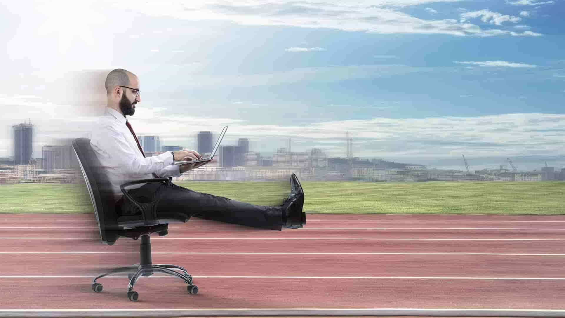 Businessman reclining in an office chair on a running track while working on a laptop, city skyline behind