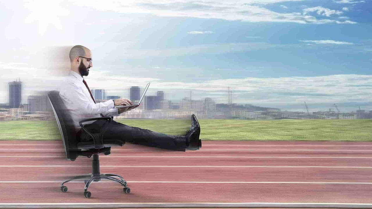 Businessman reclining in an office chair on a running track while working on a laptop, city skyline behind