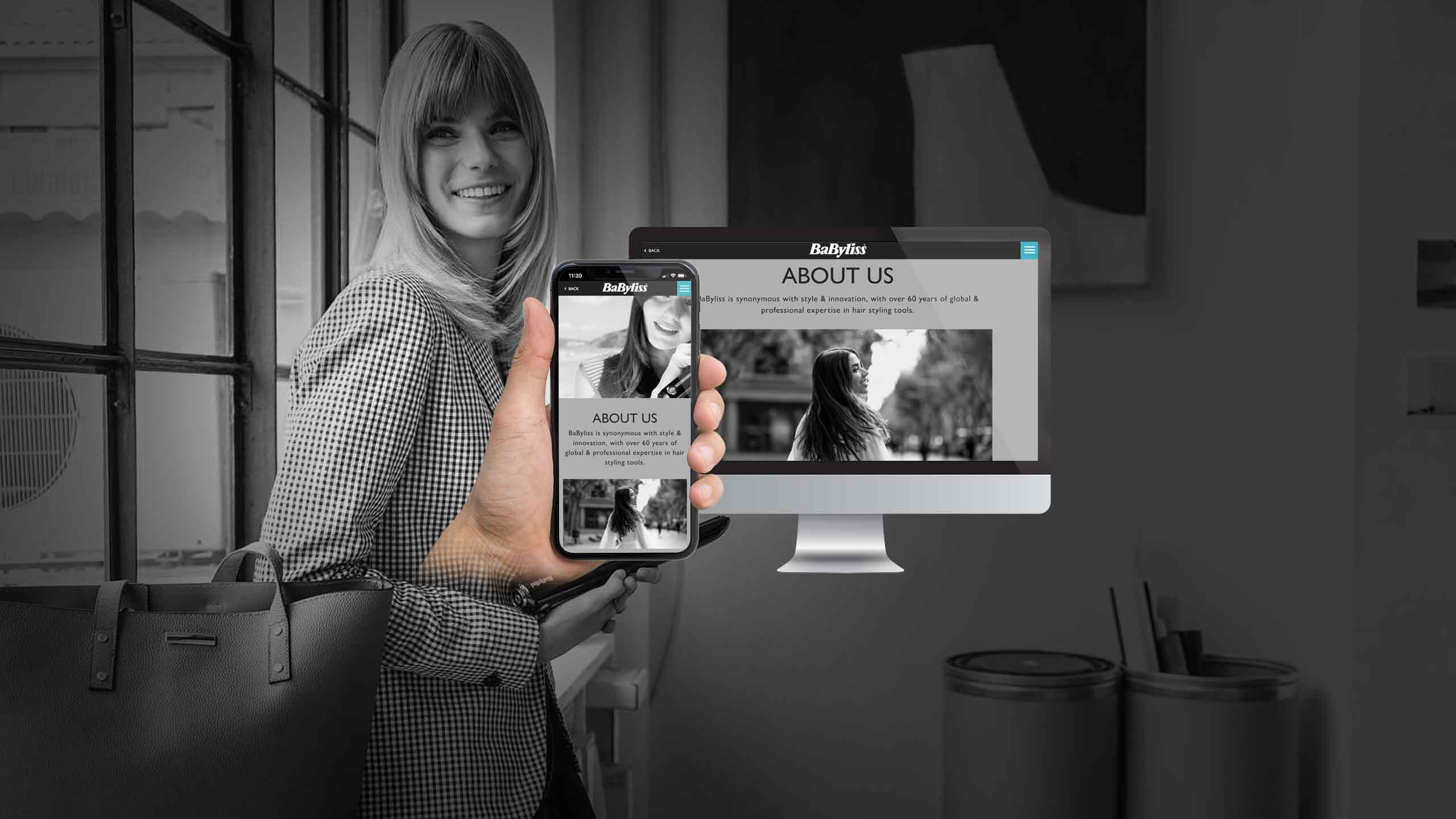Woman holding a smartphone showing the BaByliss About Us page beside a desktop monitor