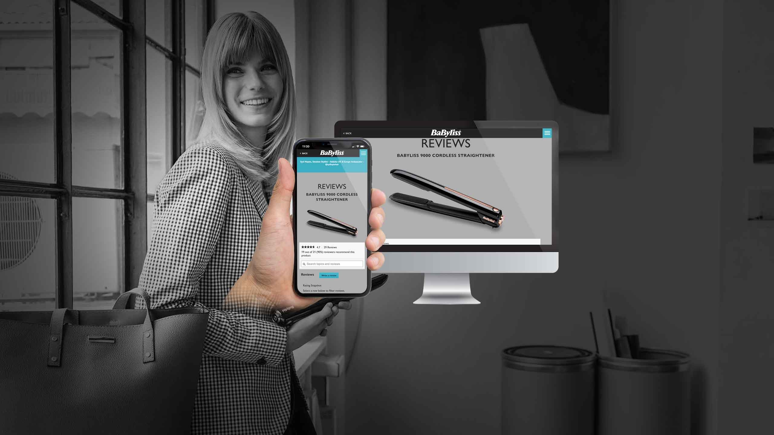 Woman holding a smartphone showing BaByliss reviews beside a desktop monitor