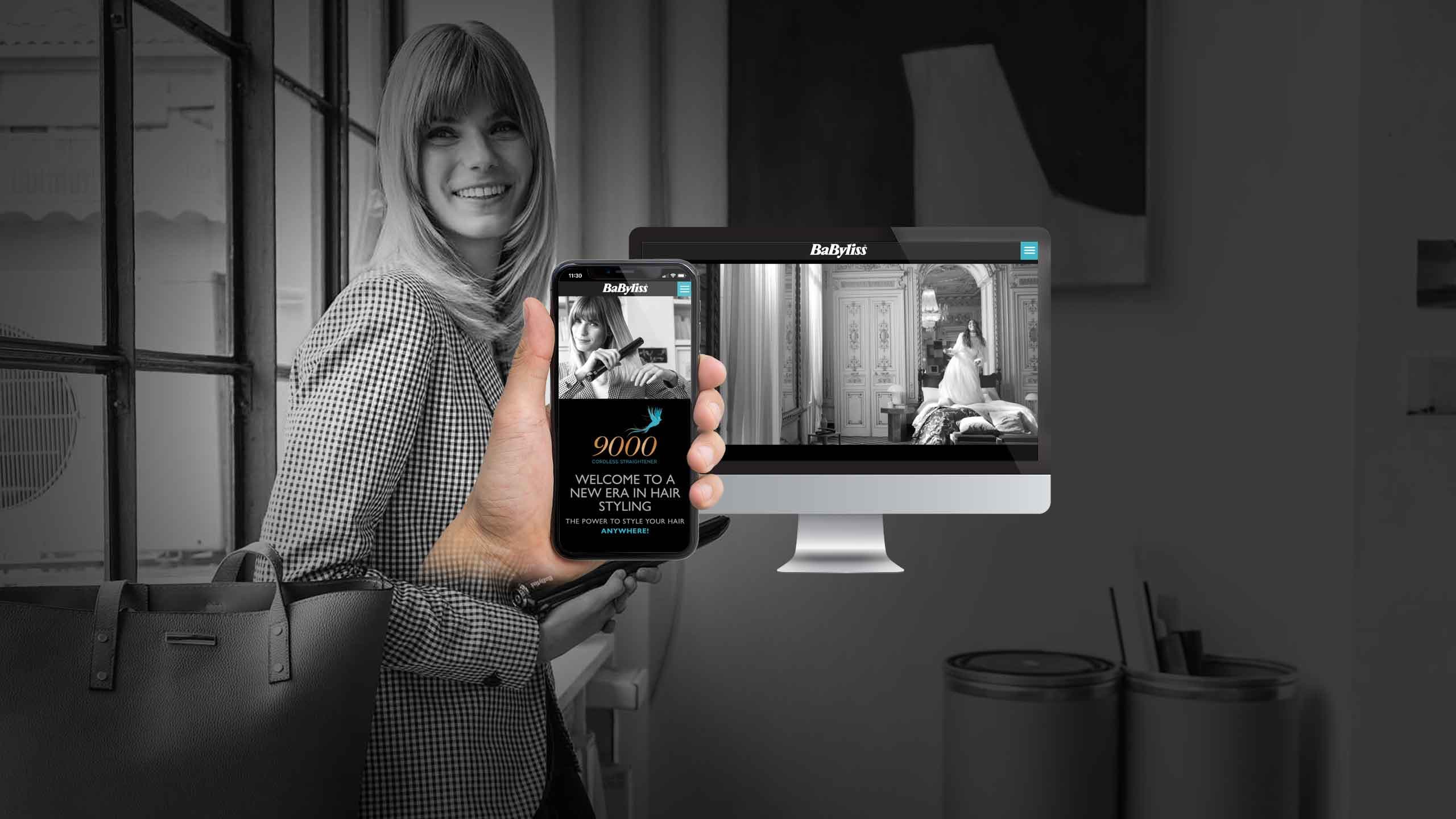 Woman holding a smartphone showing the BaByliss website beside a desktop monitor